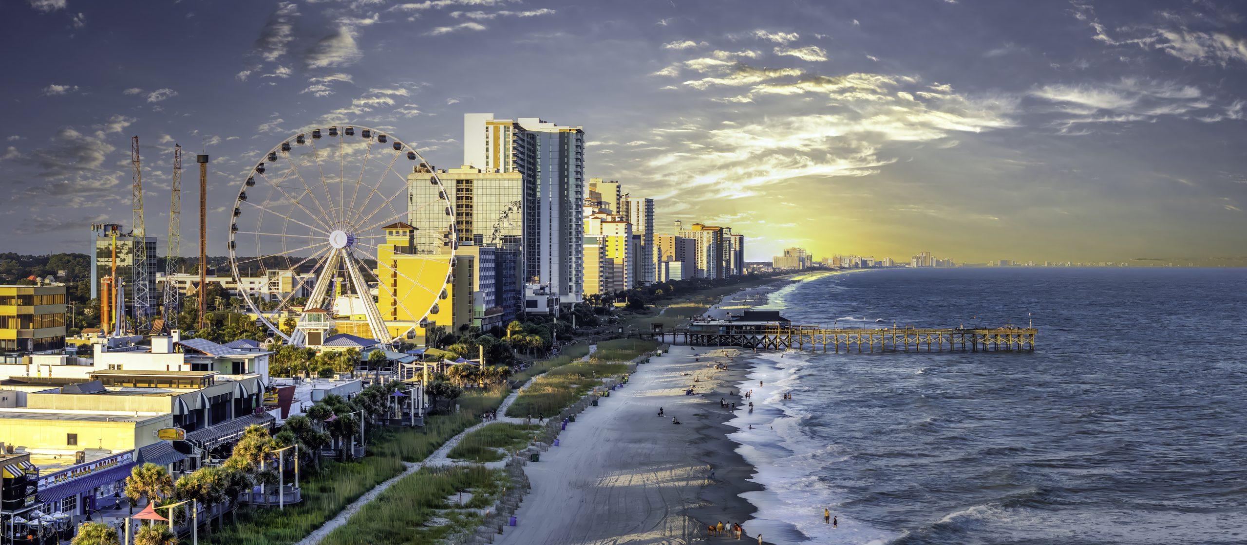 Panoramic view of Myrtle Beach skyline at sunset, featuring the SkyWheel and numerous high-rise buildings along the beachfront, with a pier extending into the ocean.